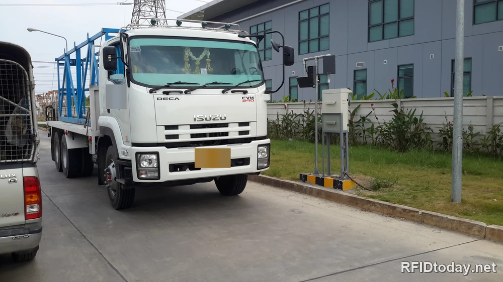 รถบรรทุกบรรทุกแร็คเหล็กกำลังขับผ่านจุดอ่าน RFID ที่ประตูโรงงาน โดยมีตู้ควบคุมและเสาเครื่องอ่านตั้งอยู่ข้างทางวิ่ง